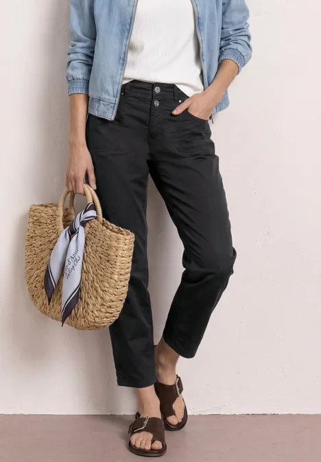 Frau trägt schwarze Hose, weißes Top, Jeansjacke, Basttasche mit Tuch und braune Sandalen.