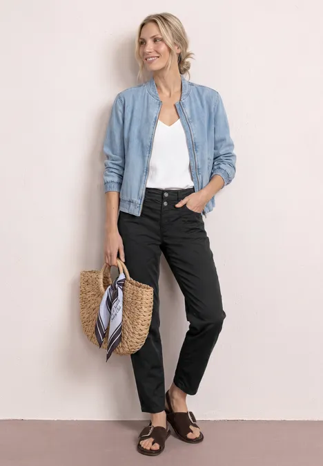 Frau in blauer Bomberjacke, weißem Top, schwarzen Jeans und Sandalen, mit Strohtasche und Tuch.