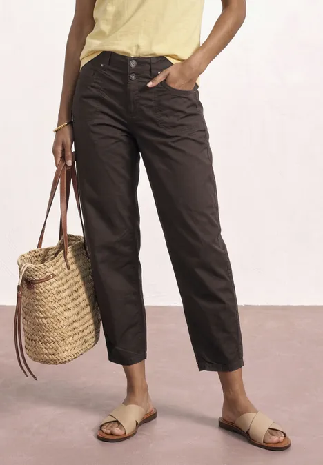 Frau trägt braune Hose, gelbes Top, geflochtene Handtasche und Sandalen.