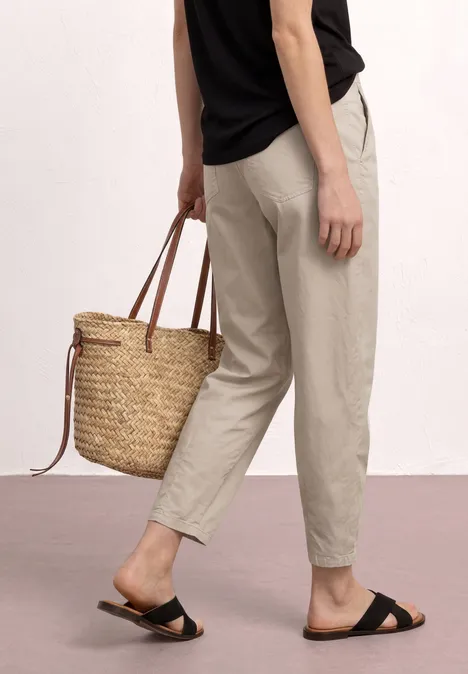Person trägt beige Hose, schwarze Sandalen und hält einen geflochtenen Strohkörben.