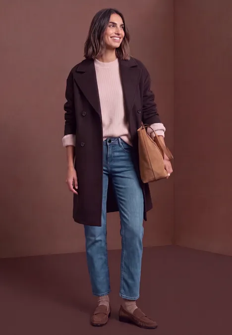 Mid Waist Straight Leg Jeans im Casual Fit Dark blue washed Eine Frau in Mantel, Pullover, Jeans und braunen Schuhen steht vor braunem Hintergrund und lächelt.