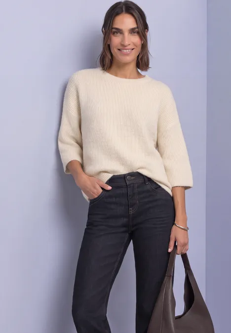 Mid Waist Straight Leg Jeans im Casual Fit Rinse Washed Frau im cremefarbenen Pullover mit 3/4-Ärmeln, schwarzen Jeans und Tasche vor neutralem Hintergrund.
