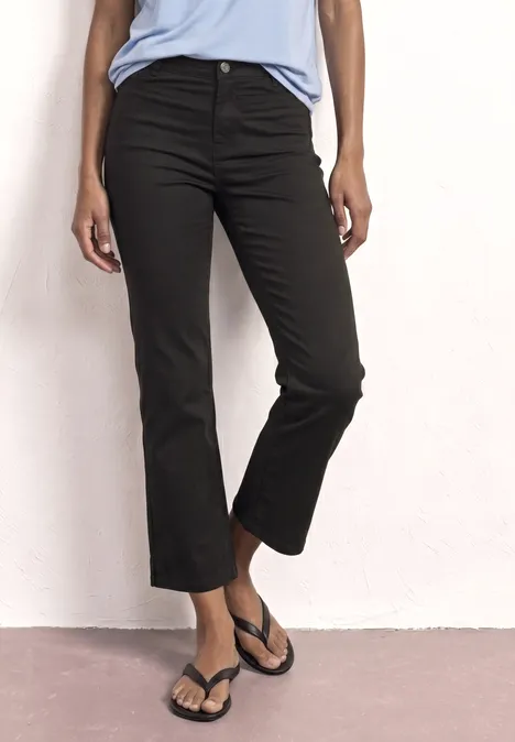 Une personne porte un pantalon noir court et évasé aux chevilles avec des tongs noires devant un mur blanc.