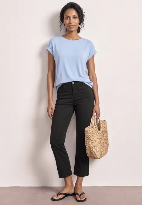 Femme devant un mur clair, portant un t-shirt bleu clair, un pantalon noir, des sandales et un sac en osier.