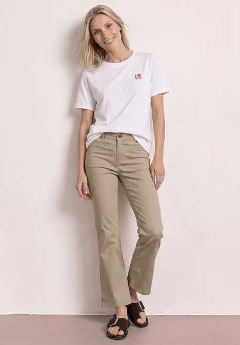 Femme appuyée sur un mur clair, porte un t-shirt blanc, un pantalon beige ample 7/8 et des sandales noires.