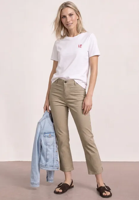 Une femme porte un T-shirt blanc avec un petit cœur rouge, un pantalon beige, des sandales noires et tient une veste en jean.