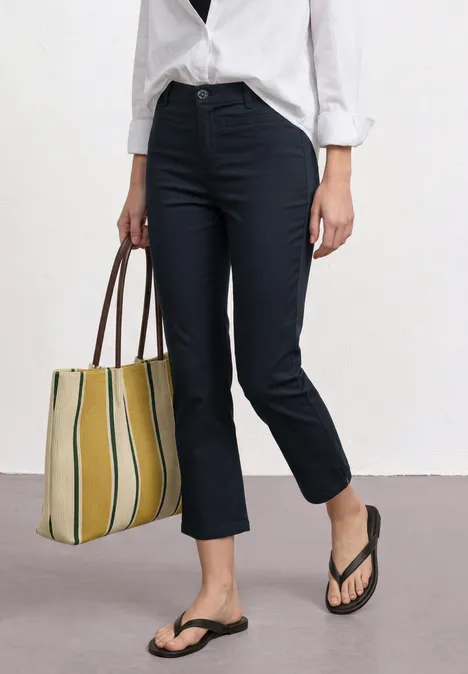 3/4 broek met flared pijpen in casual pasvorm shadowed navy Vrouw in donkerblauwe, hoge taille broek, wit overhemd, geel-wit gestreepte tas en zwarte slippers.