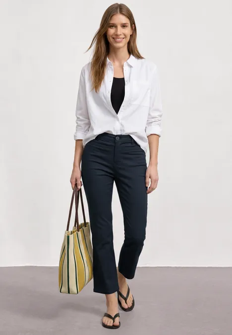 3/4 broek met flared pijpen in casual pasvorm shadowed navy Vrouw met witte geknoopte blouse, zwart topje, donkere broek, slippers en gestreepte tas.