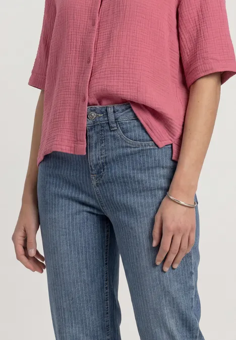 Eine Person trägt eine rosafarbene, kurzärmelige Bluse, blaue Jeans und ein Armband, nur Oberkörper und Hände sichtbar.