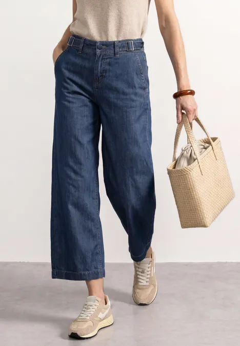 Frau trägt weite, knöchellange Jeans, Sneakers und hält eine geflochtene Korbtasche.