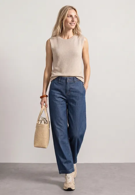 Eine Frau in beigem Top, weitem blauen Jeans und Sneakers, hält eine Basttasche vor hellem Hintergrund.