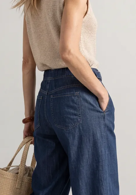 Frau von hinten in weitem Jeans und beigem Top, hält eine Strohtasche.