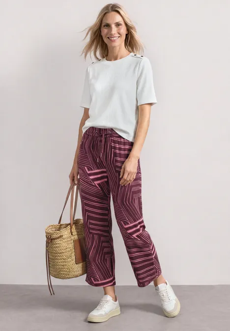 Culotte taille haute à motifs blush berry Frau in weißem T-Shirt, gemusterten lila Hosen, weißen Sneakern, hält Korbtasche vor neutralem Hintergrund.