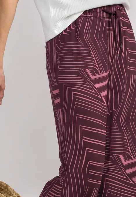 Culotte taille haute à motifs blush berry Eine Person trägt eine weiße Bluse und weinrote Hose mit pinkem, geometrischem Muster.