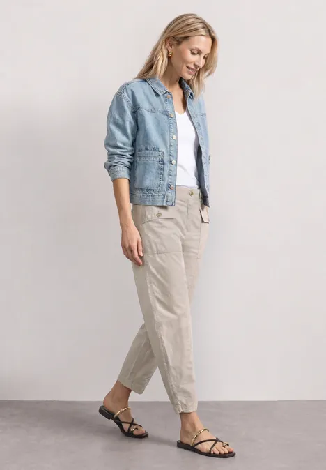 Frau in hellblauer Jeansjacke, weißem Top und beigen Stoffhosen mit schwarzen Riemchensandalen vor hellem Hintergrund.