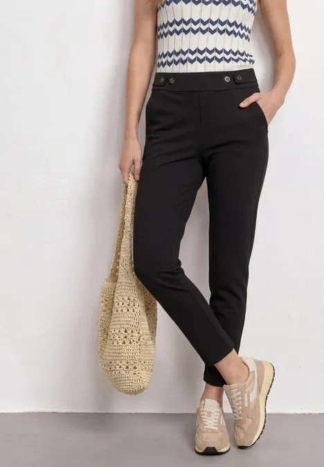 Frau in schwarzen Slacks, gemustertem Top, trägt Strohbeutel und bequeme beige Sneaker.