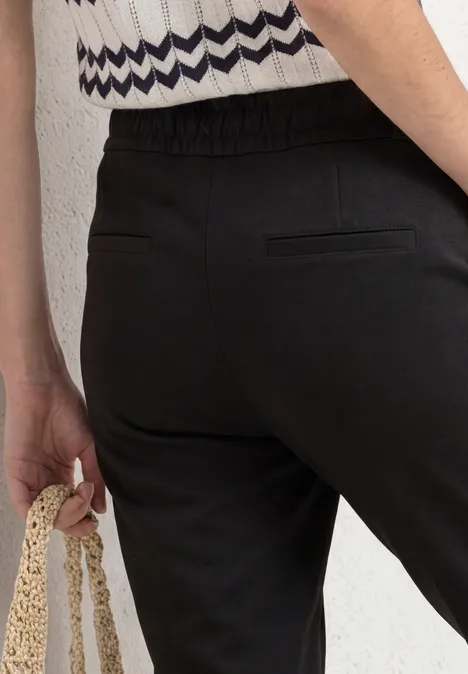 Nahaufnahme einer Person in schwarzer Hose mit elastischem Bund und Gesäßtasche, die eine gehäkelte Tasche hält.