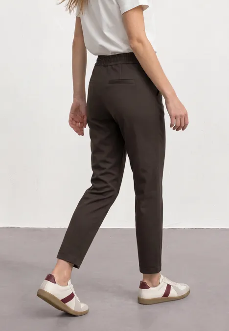 Frau trägt schmale, dunkelbraune Hose und weiße Sneaker mit rotem V-Design vor heller Wand.