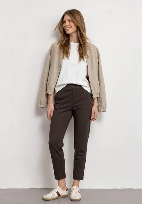 Eine junge Frau in weißem Shirt, beigem Blazer, dunklen Hosen und Sneakers vor hellgrauem Hintergrund.