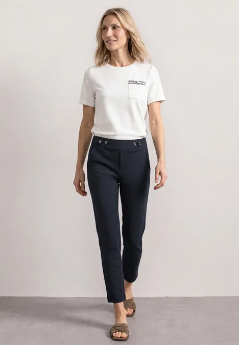 Frau mit weißem Shirt, dunkelblauen knöchellangen Hose und Sandalen vor neutralem Hintergrund