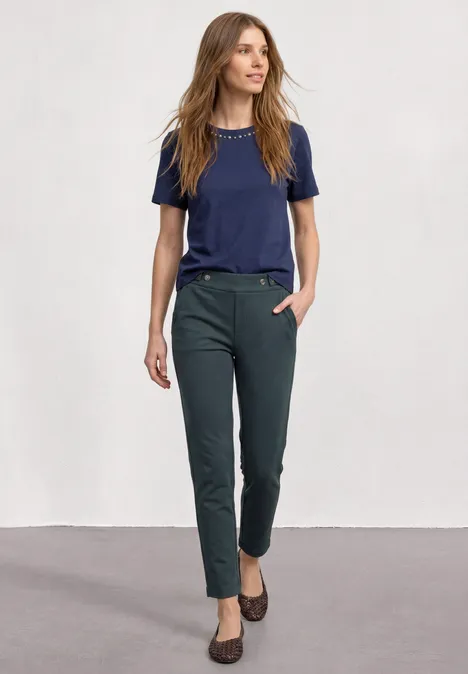 Frau in dunkelgrünen, schmalen Hosen, marineblauem T-Shirt und braunen Schuhen vor hellem Hintergrund.