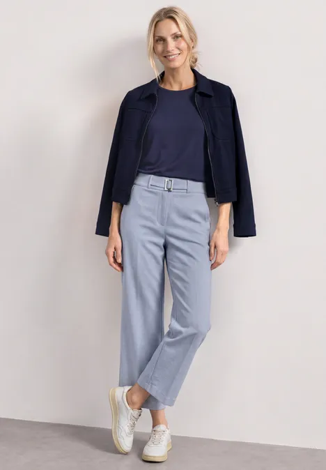 High Waist Culotte met gespdetail whispery blue Een vrouw in een marineblauw topje en jasje, lichtblauwe broek en witte sneakers tegen een neutrale achtergrond.