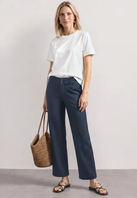 Frau in weißem T-Shirt, dunkelblauen Jeans, schwarzen Sandalen und Strohtasche vor neutralem Hintergrund.