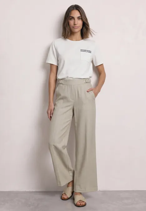 High Waist Straight Leg Hose im Casual Fit dark creamed sand Frau in weißem T-Shirt, beiger, weiter Hose und Sandalen vor neutralem Hintergrund.