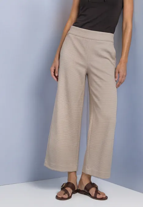 High Waist Culotte im Casual Fit saddle brown Frau in beigen, weit geschnittenen 7/8-Hosen und braunen Sandalen vor neutralem Hintergrund.