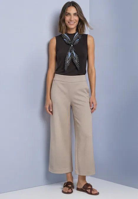 High Waist Culotte im Casual Fit saddle brown Frau in schwarzem Top, gemustertem Halstuch, beiger Culotte und braunen Sandalen vor blauem Hintergrund.
