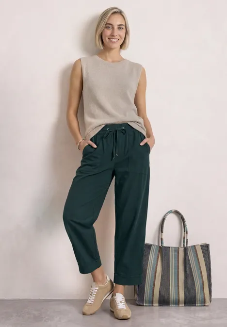 Femme souriante en débardeur beige, pantalon large noir, baskets et grand cabas rayé sur fond clair.