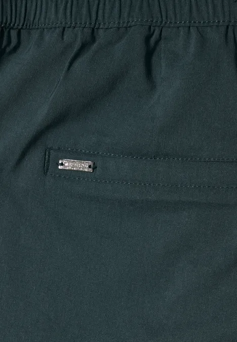 Gros plan d’un pantalon sombre avec poche zippée et petit logo métallique.