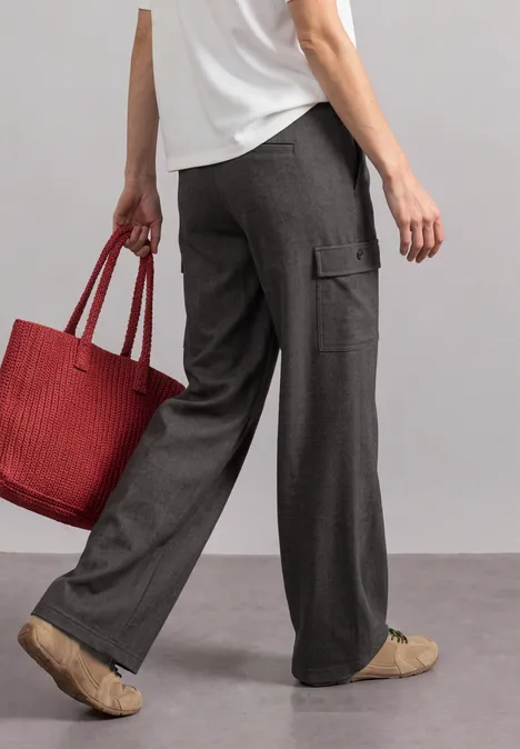 Vrouw in wijde grijze stofbroek, wit shirt en beige sneakers, met een grote rode shopper in de hand.