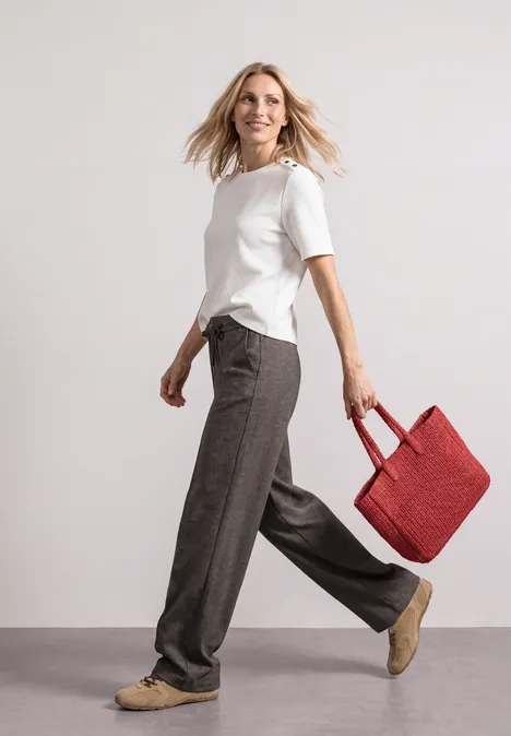 Vrouw in wit T-shirt en grijze broek loopt vrolijk met een rode tas in haar hand.
