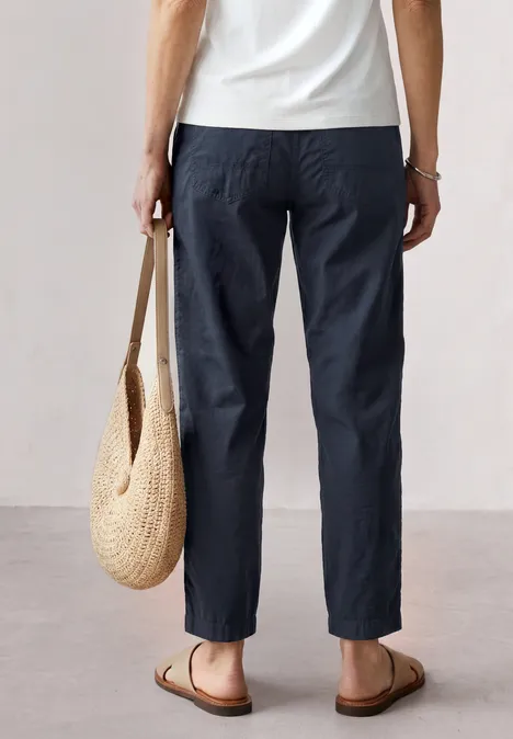 Rückansicht einer Person in dunkler Hose, weißem Oberteil, Sandalen und geflochtener Strandtasche.