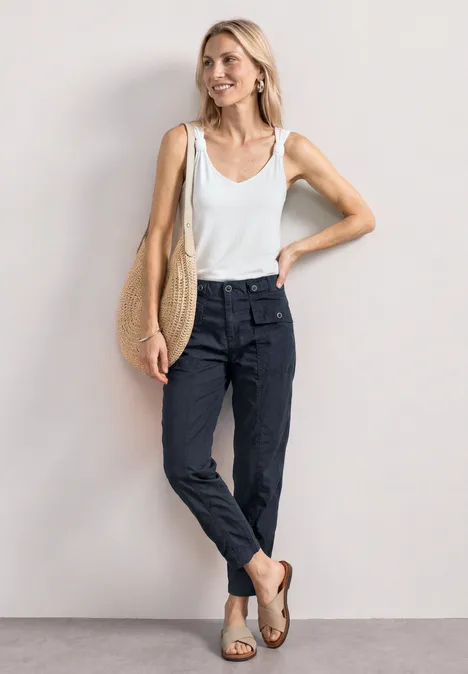 Frau in ärmellosem weißem Top, dunklen Bundfaltenhosen, Sandaletten und Stofftasche, lässig an die Wand gelehnt
