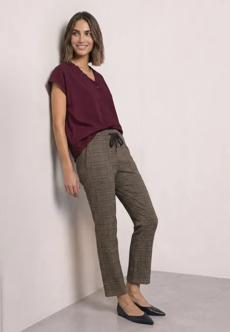 Eine Frau lehnt an einer Wand, trägt bordeauxrotes Oberteil, karierte Hose und schwarze Ballerinas.