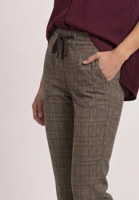 Eine Person trägt karierte, hoch taillierte Stoffhose mit Kordelzug und Eingrifftaschen in Braun.