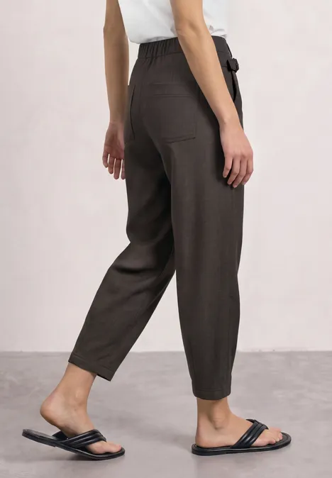 High Waist Barrel Leg Hose im Casual Fit black coffee Frau in hoch geschnittenen schwarzen Stoffhosen mit Taillengürtel und schwarzen Sandalen vor neutralem Hintergrund.