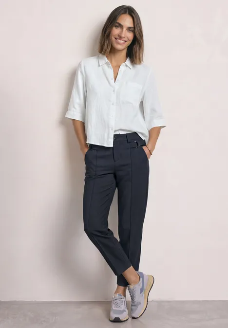 High Waist Barrel Leg Hose im Casual Fit darkest blue Eine lächelnde Frau in weißer Bluse, dunkler Stoffhose und Sneakern vor hellem Hintergrund.