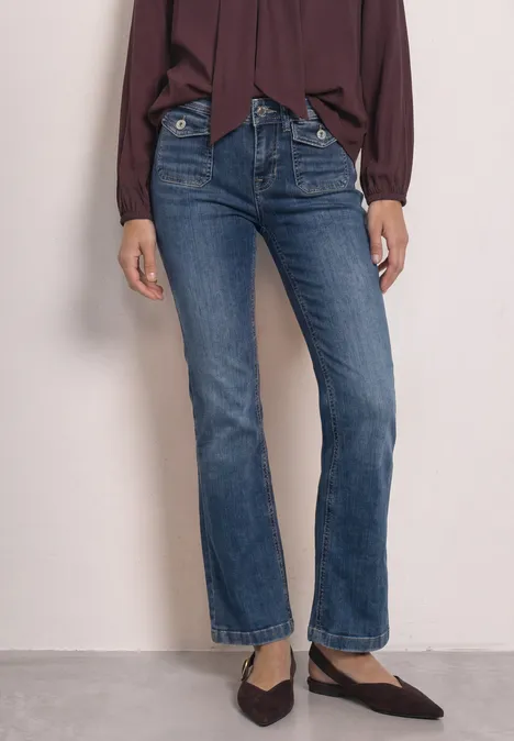 Mid Waist Bootcut Jeans im Slim Fit Medium Blue Random Washed Frau in dunkelblauen, geraden Jeans und spitzen Ballerinas, kombiniert mit einer weiten, dunkelroten Bluse.