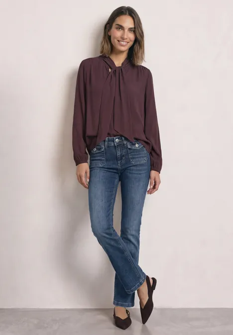 Mid Waist Bootcut Jeans im Slim Fit Medium Blue Random Washed Frau in bordeauxrotem Blusenshirt und blauen Bootcut-Jeans steht lächelnd vor hellem Hintergrund.