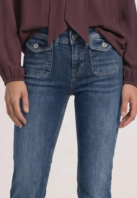 Mid Waist Bootcut Jeans im Slim Fit Medium Blue Random Washed Nahaufnahme einer Person in dunkelblauen Jeans mit großen vorderen Hosentaschen und roter Bluse