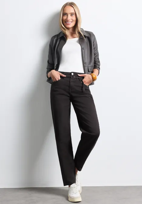 High Waist Barrel Leg Jeans im Loose Fit Brown Washed Eine junge Frau in schwarzer Jeans, weißem Shirt und grauer Jacke steht lächelnd vor hellem Hintergrund.