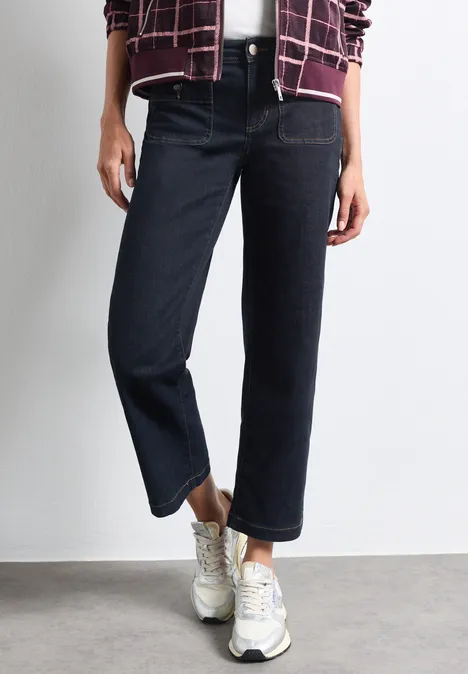 Persoon draagt donkere, rechte jeans en witte sneakers, staand op een grijze vloer.