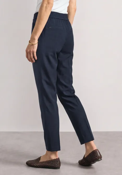 Person trägt dunkelblaue, gerade geschnittene Hose mit elastischem Bund und geflochtenen Loafers.
