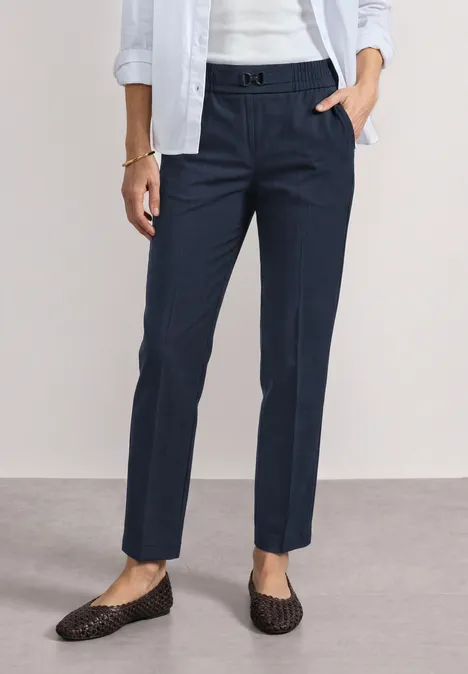 Dame in dunkelblauen Chino-Hosen mit Bügelfalte trägt weiße Bluse und geflochtene schwarze Loafers.