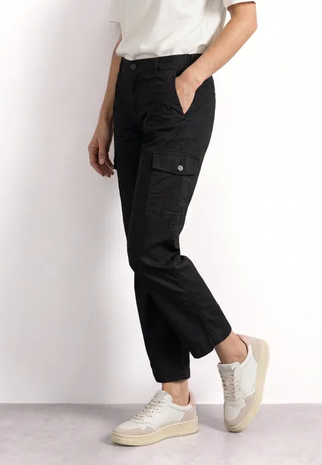 Une personne porte un pantalon cargo noir et des baskets blanches sur fond neutre.