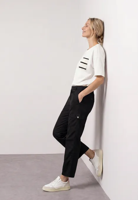 Femme appuyée contre un mur blanc, porte un t-shirt blanc, un pantalon cargo noir et des baskets blanches.