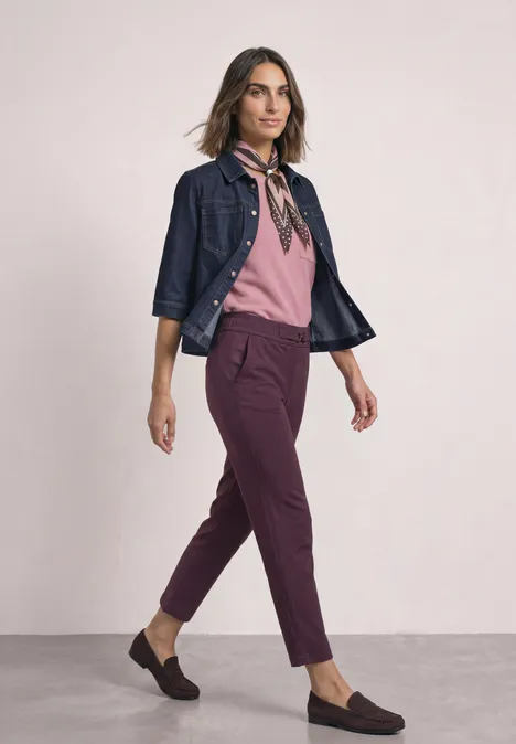 Vrouw in stijlvolle look: roze blouse, bordeaux broek, spijkerjack, sjaal en zwarte loafers.